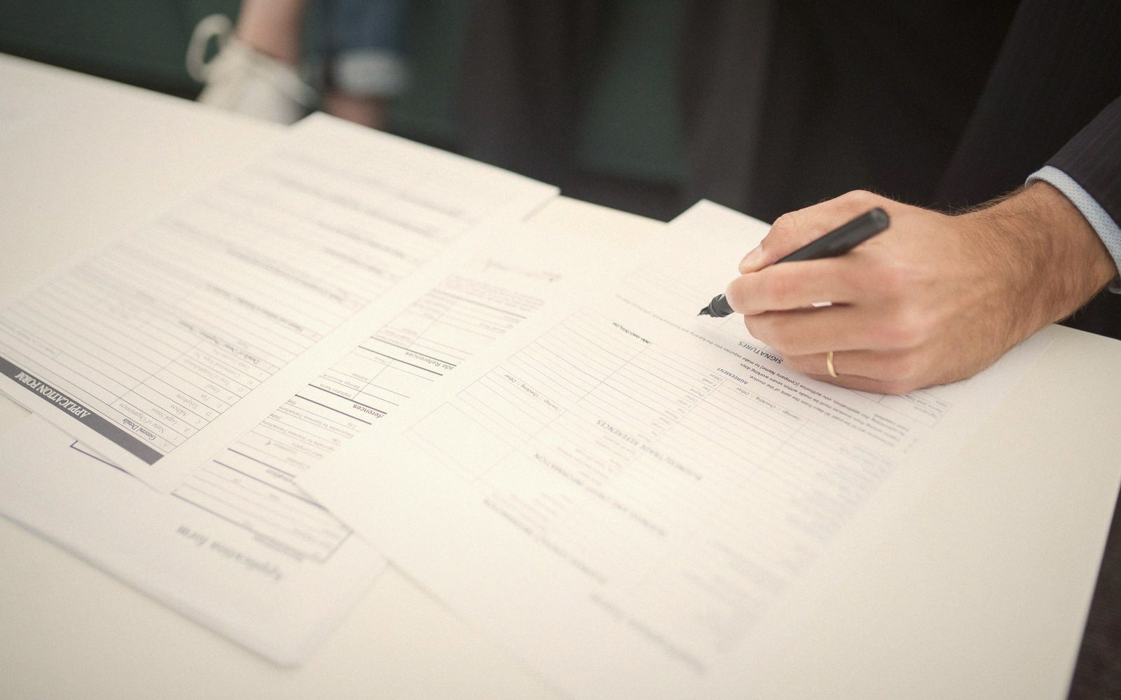 A hand in a suit sleeve signs a printed form with a black pen