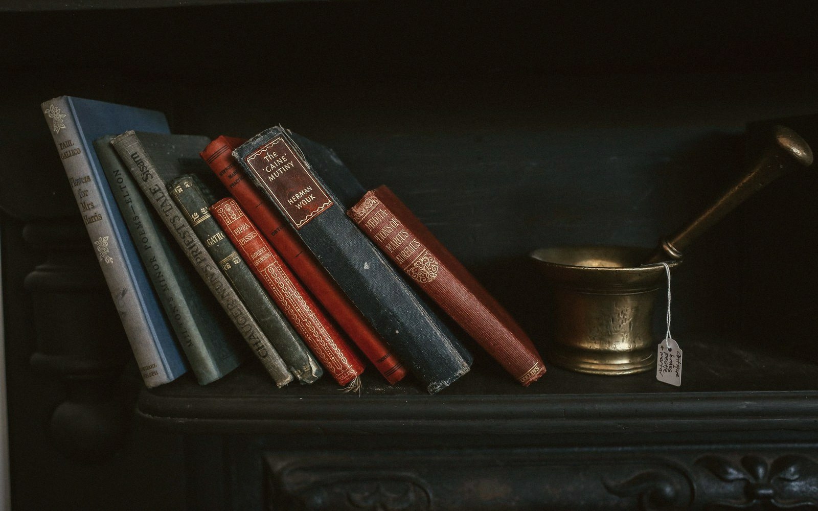 Old hardback books leaning on a mantelpiece beside a brass mortar and pestle with a price tag